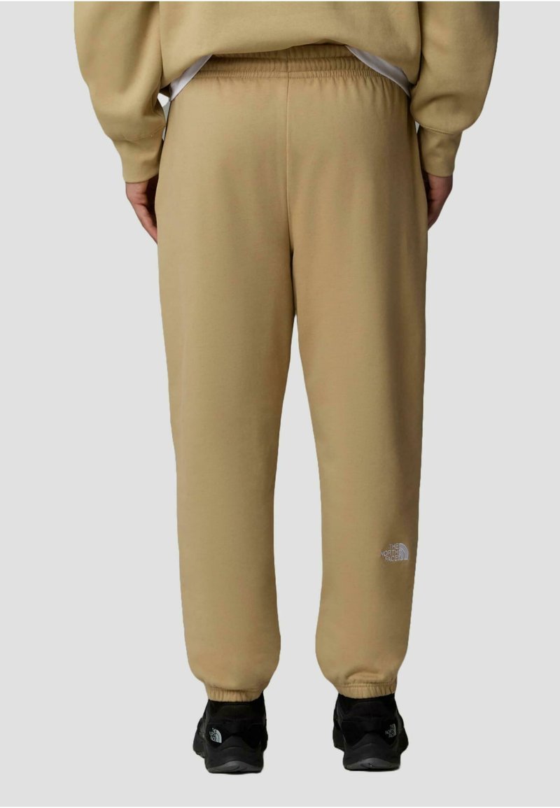 Pantalons de survêtement beige en matériau doux, avec une coupe décontractée et une taille élastique. Le logo The North Face est imprimé en blanc sur la jambe inférieure.