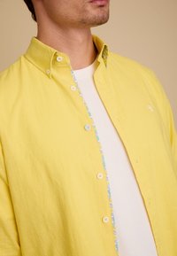 Geel overhemd met knopen gemaakt van een geweven stof, met een patroon aan de binnenvoering en witte knopen, in lagen gedragen over een wit t-shirt.