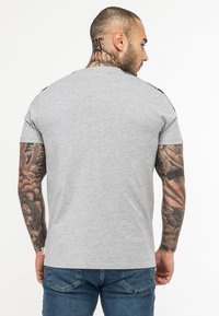 Tricou gri cu mâneci scurte, decolteu rotund și accente negre texturate pe umeri. Model îmbrăcat cu blugi din denim; tatuaje vizibile pe brațe.