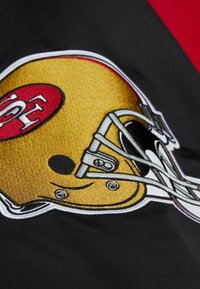 Gesticktes Football-Helm-Design auf schwarzem Stoff, mit einem glänzenden goldenen Helm, rotem Logo und weißen Akzenten, das komplizierte Details zeigt.