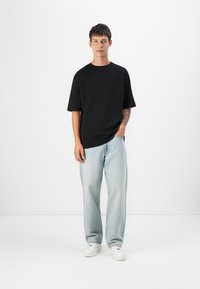 Sort bomulds T-shirt med korte ærmer og afslappet pasform, båret med lyseblå posede jeans og hvide sneakers. Baggrunden er simpel hvid.