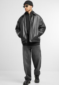 Veste bomber en cuir noir sur un pull en laine noir, assortie à un pantalon gris ample et des bottes noires. Les accessoires incluent des lunettes de soleil vertes et un bonnet noir.