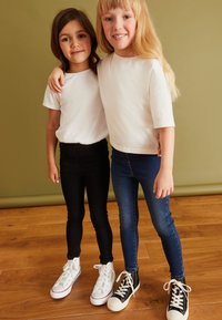 To børn står side om side iført hvide t-shirts, sorte skinny jeans og blå jeans. De har hvide og sorte sneakers på.