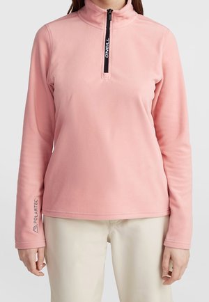 Woman wears light pink long-sleeve fleece pullover with black half-zip and beige pants, standing against a plain background.