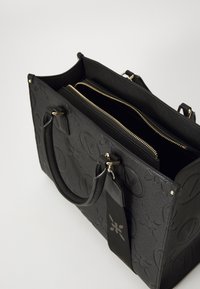 Svart teksturert totebag med pregete mønstre, doble håndtak og gullfargede glidelåslommer. Har et bagasjelapp-akcent.