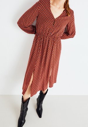 Femme portant une robe midi rouge à motifs avec manches longues, fentes devant, et des bottes de cowboy noires à bout pointu, debout devant un fond uni.