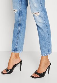 Mule nere con tacco, punta aperta e fronte quadrato, caratterizzate da una texture liscia in pelle. Indossate con jeans azzurri chiari consumati.