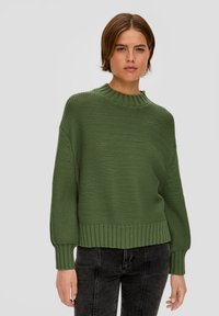 Vrouw in een overmaatse olijfgroene gebreide trui en zwarte gewassen jeans, staande tegen een eenvoudige lichtgrijze achtergrond.