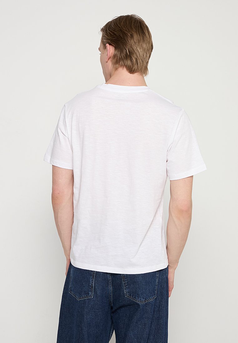 Camiseta blanca de algodón con cuello redondo, vista desde atrás, con mangas cortas y un corte relajado, combinada con jeans oscuros de mezclilla.