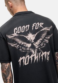 Zwarte T-shirt met korte mouwen, voorzien van een grote afbeelding van een gevleugeld figuur en vetgedrukte tekst "GOOD FOR NOTHING" in contrasterende lichte kleuren.