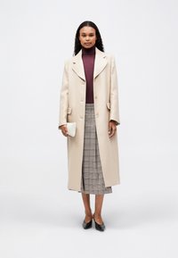 Ženska stoji oblečena v bež dolg plašč, burgundski pulover s turtleneck ovratnikom, pikčasto krilo, črne čevlje in drži majhno belo pleteno torbico.