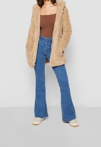 Person iført sandfarvet teddyfrakke, brun top med firkantet halsudskæring, blå høj talje flared jeans og beige sneakers står på gråt gulv.