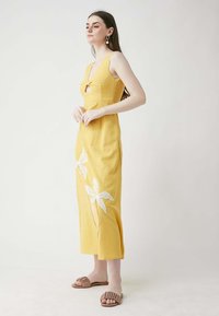 Gelbes Leinenkleid mit tiefem V-Ausschnitt und Schlüssel-Ausschnitt, verziert mit weißen gestickten Blumen am Saum. Das Modell trägt flache Sandalen.