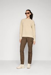 Beige sweater met wafelstructuur, bruine cargobroek met zijzak en lichtgekleurde sneakers. Model draagt een zonnebril met bruine glazen.