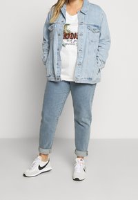 Ljusblå denimjacka över en vit T-shirt med tryckt design, i kombination med ljusblå jeans och vita sneakers med svarta detaljer.