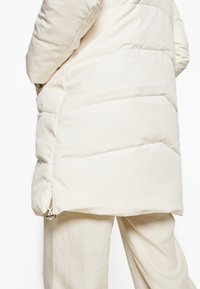 Giacca a vento bianca con design trapuntato e tasche laterali con zip. Presenta un orlo regolabile con coulisse e una texture di stoffa liscia. Indossata sopra pantaloni di colore chiaro.