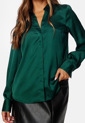 Vrouw draagt een donkergroene satijnen blouse met lange mouwen en een zwarte leren rok, hand in de zak van de rok.