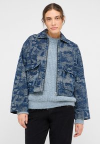 Veste en denim bleu coupée avec une texture à motif, fermeture éclair et deux poches avant, portée sur un pull en maille bleu clair.