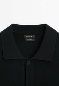 Polo en maille noire avec un col pointu et un bouton visible, montrant l’étiquette de la marque au niveau de l’encolure.