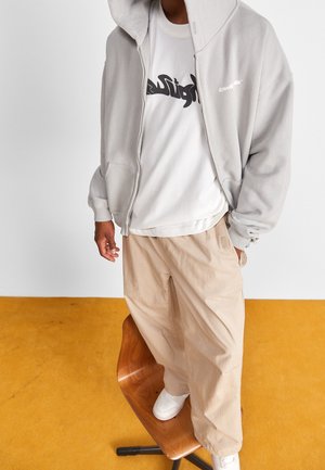 Grijze zip-up hoodie over een wit t-shirt met een zwart grafisch logo, gecombineerd met beige wijde broek en witte sneakers, staand op een stoel.