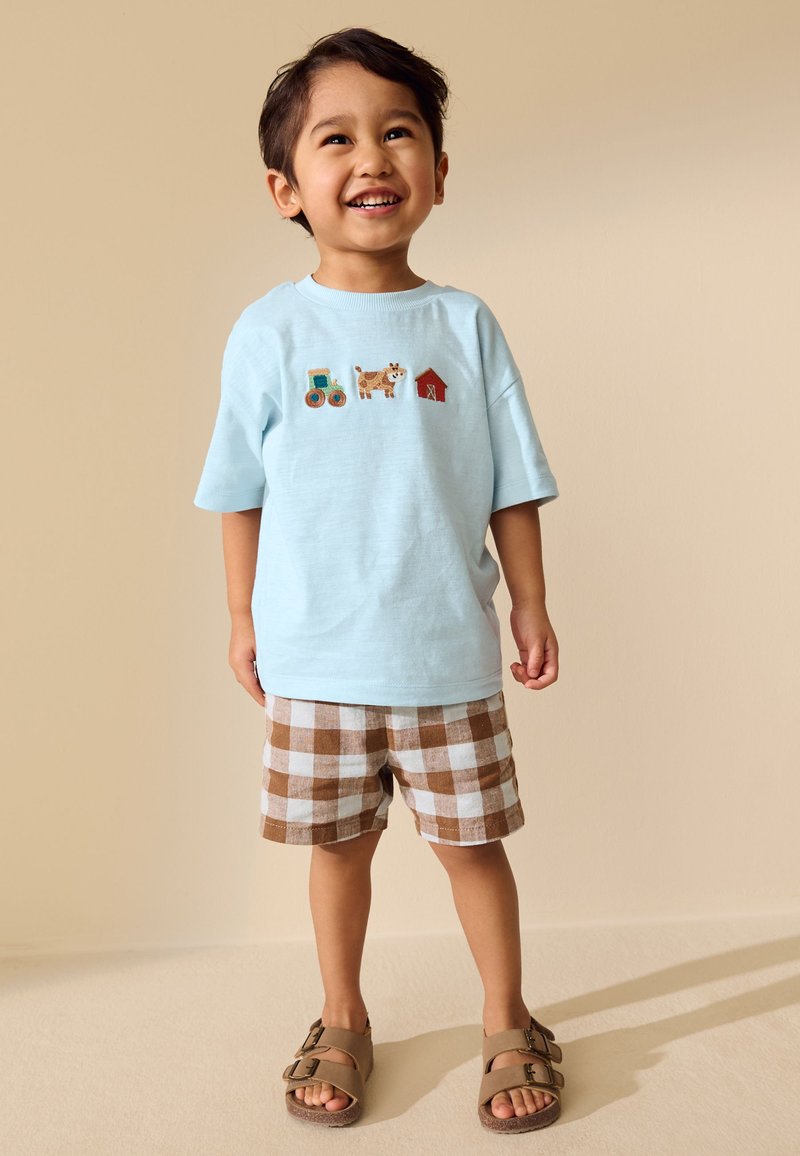 Joven sonriente con una camisa azul clara con diseños de tractor, vaca y granero, pantalones cortos marrones a cuadros y sandalias color topo, de pie en el interior.