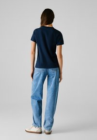 T-shirt navy a maniche corte abbinato a jeans a vita alta larghi color blu chiaro. Il modello indossa sneaker color crema, mostrando un look casual.