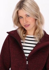 Burgundska zip-up jakna z teksturiranim pletenim zaključkom, z visokim ovratnikom in kontrastno črno-belo črtasto srajco spodaj.