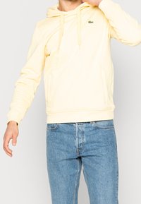 Person som bär en ljusgul hoodie med dragsnören och en liten grön krokodil-logotyp, kombinerad med blå denimjeans, stående mot en enfärgad bakgrund.