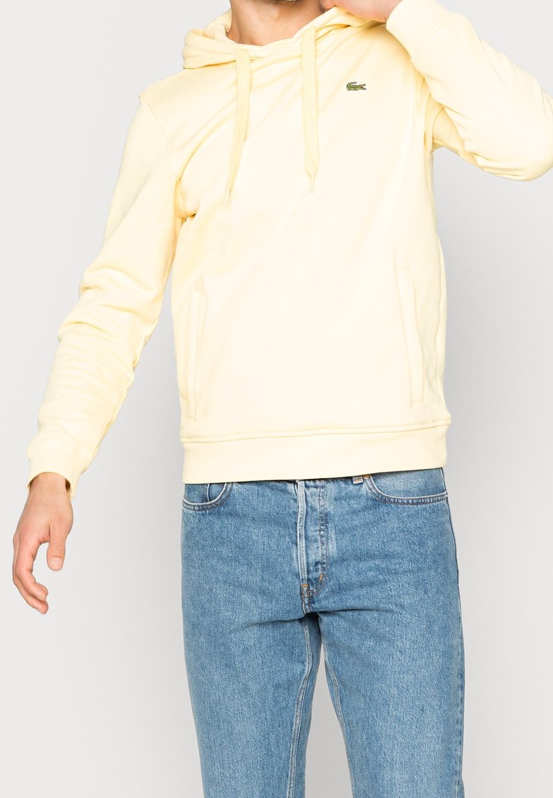 Person som bär en ljusgul hoodie med dragsnören och en liten grön krokodil-logotyp, kombinerad med blå denimjeans, stående mot en enfärgad bakgrund.