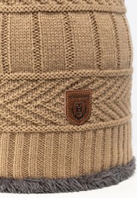 Bonnet tricoté beige avec une texture côtelée, présentant un patch en cuir marron embossé d'un lion et "JEANSCODE". Bord en fausse fourrure gris au bas.