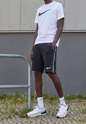 Osoba nosząca białą koszulkę Nike, czarne krótkie spodenki Nike, białe skarpetki Nike i białe buty sportowe Nike stoi na brukowanej nawierzchni obok metalowej poręczy.