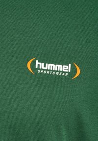 Zaļš Hummel sporta t-krekls ar gludu, teksturētu audumu, kas rotāts ar baltiem un oranžiem logotipa akcentiem, un klasisku apaļu kakla izgriezumu.