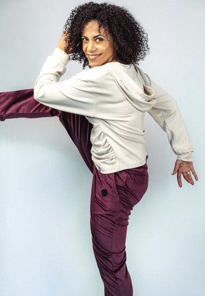 Kohar juustega naine, kes kannab beežikat kapuutsiga pusa ja lillaid pükse, tõstab ühe jala kõrgele, naeratades ühevärvilise tausta ees.