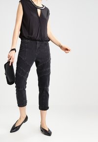 Haut sans manches noir avec un décolleté en trou de serrure, associé à un pantalon noir fuselé avec des détails en panneaux. Le mannequin tient une pochette noire et porte des ballerines noires.