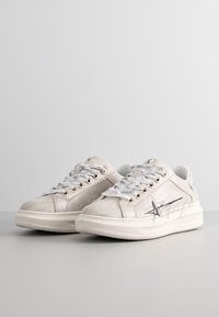 Ljust grå mocka sneakers med silverdetaljer, rund tå, platta snören och grafiska designdetaljer på sidan.