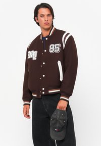 Uomo che indossa una giacca varsity marrone con strisce bianche e toppe, tiene un cappellino da baseball nero nella mano destra.