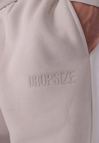 Világosszürke vászonból készült melegítőnadrág, melyen a dombornyomott "DROPSIZE" felirat látható. Az anyag puha és rugalmas, jól látható varrási részletekkel.