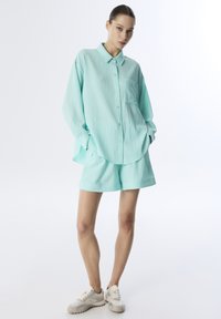 Camisa button-up de color verde menta con textura y un bolsillo en el pecho, combinada con pantalones cortos a juego. Lleva zapatillas claras, mostrando un ajuste relajado.
