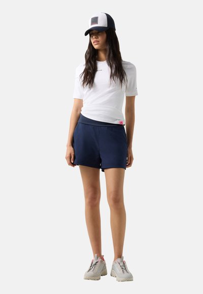 Frau trägt weißes T-Shirt, dunkelblaue Shorts, weiße Sneakers und eine schwarz-weiße Kappe, steht mit entspannt an den Seiten hängenden Händen.