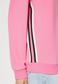 Sudadera rosa con puños y dobladillo acanalados, con franjas laterales en negro y blanco. Textura suave, corte casual y tejido blando.