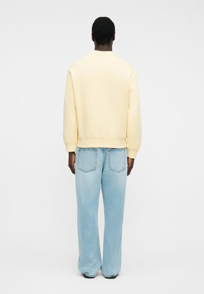 Άνδρας που φοράει ανοιχτό κίτρινο φούτερ και ανοιχτό μπλε τζιν παντελόνι με φαρδιά μπατζάκια, στέκεται γυρισμένος προς τα πίσω μπροστά σε λευκό, απλό φόντο.