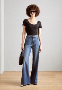 Svart kortärmad bodysuit, högmidjade blå denimbyxor med utsvängda ben och svarta solglasögon. Modellen bär en svart handväska. Trägolvsbakgrund.