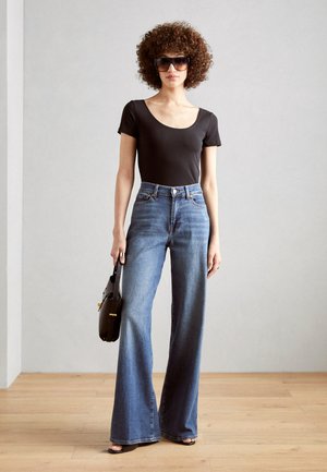 Svart kortermet bodysuit, høyreiste blå denim flared jeans og svarte solbriller. Modellen bærer en svart håndveske. Bakgrunn av tregulv.