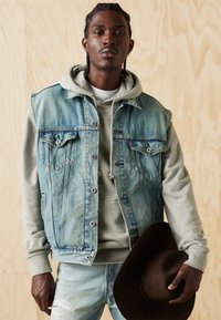 Mėlyno plovimo džinsinė liemenė be rankovių, dėvima virš pilkos gaubtuotos megztinės. Šviesiai mėlynos, dėvėtos kelnės iš džinso audinio. Modelis laiko rudos spalvos kepurę. Medinių lentų fonas.