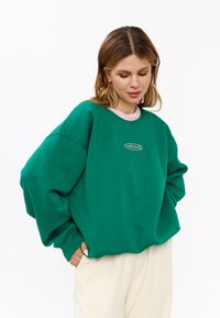 Felpa oversize verde in cotone, con collo rotondo, polsini a costine e un delicato logo ricamato sul petto.