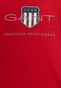 Camiseta roja con un logo de escudo con rayas verticales en marrón y blanco, y texto que dice "GANT 1949 AMERICAN SPORTSWEAR" en gris.