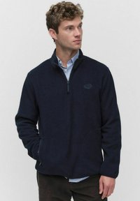 Granatowy polar z półzamkiem w kołnierzu, przednimi kieszeniami i miękką teksturą. Nosi się go na koszuli z kołnierzykiem, z detalem logo.