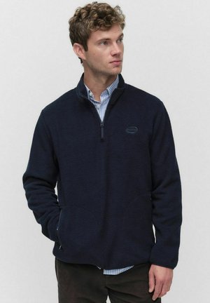 Marinblå fleece-pullover med halvdragkedja, framfickor och mjuk struktur. Bärs över en skjorta med krage och har detalj med logotyp.