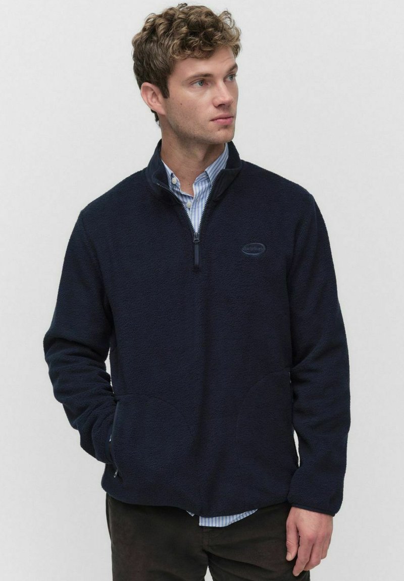 Granatowy polar z półzamkiem w kołnierzu, przednimi kieszeniami i miękką teksturą. Nosi się go na koszuli z kołnierzykiem, z detalem logo.