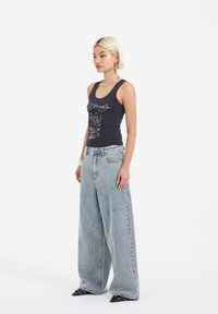 Czarny top bez rękawów z graficznym wzorem, w połączeniu z oversize'owymi, jasnoniebieskimi dżinsami. Model nosi spiczaste czarne buty i srebrną biżuterię.